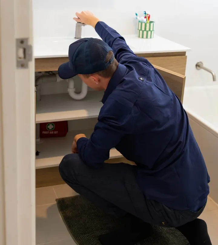 Plumber in a cap crouched at an open bathroom vanity checking the waste pipe under the basin