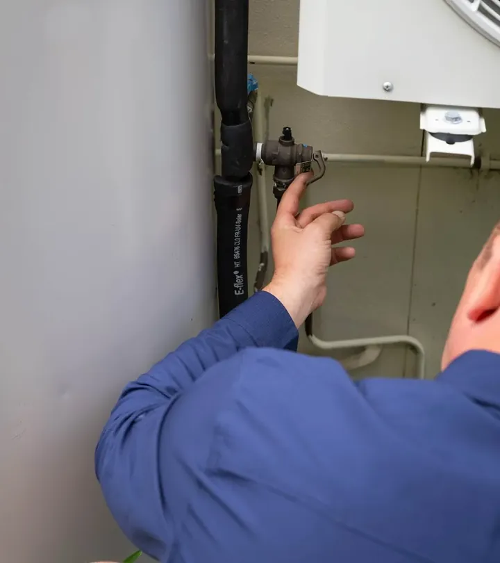 Plumber's hand on the relief valve and hose fittings beside a hot water tank