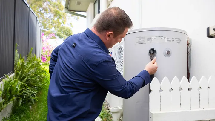 Plumber in a navy shirt adjusting the valve on a hot water system mounted on an outside wall
