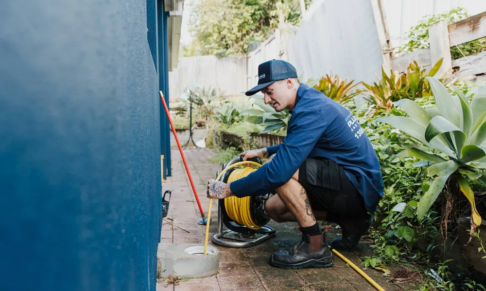 Plumber crouched beside a drain cleaning machine on a paved path next to a house
