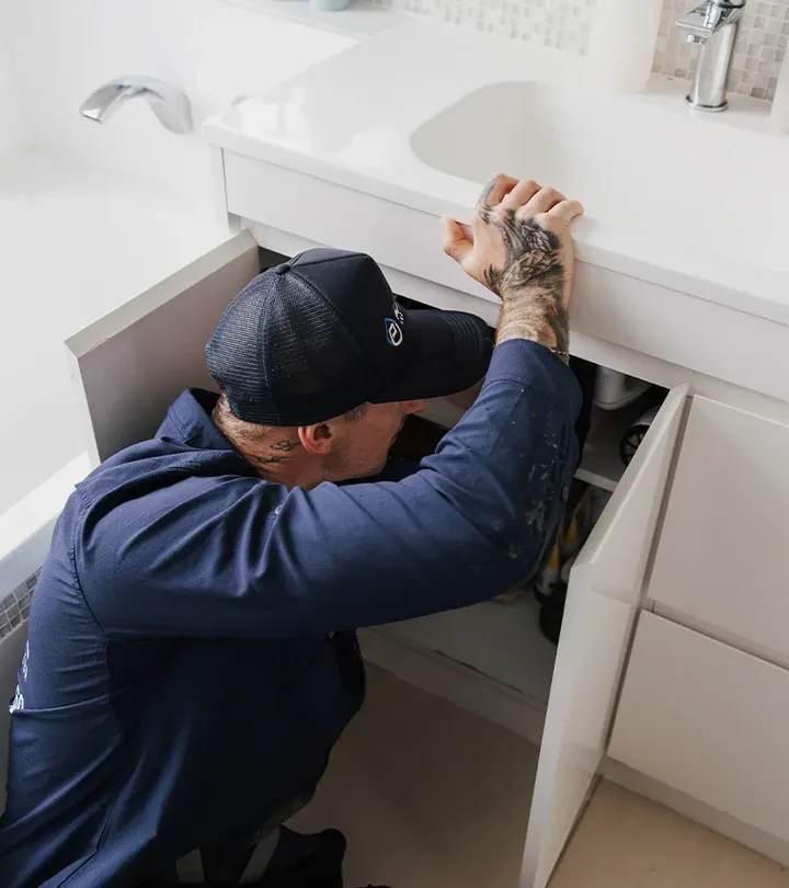 Plumber in a cap reaching into the cupboard under a bathroom vanity next to a bath